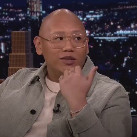 Jacob Batalon is wearing glasses, gold chain and ring, and grey jacket.